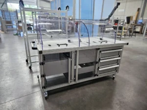 Banc de test mobile en profilé aluminium avec capots de protection polycarbonate relevés, plateau usiné, tiroirs latéraux et bras support écran, sur roulettes pivotantes
